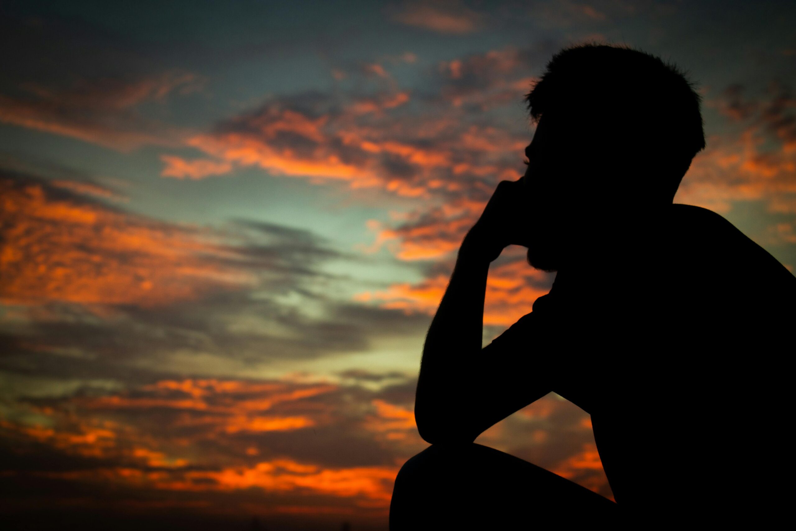 A silhouette of a man sitting and contemplating against a vibrant sunset sky filled with warm hues.